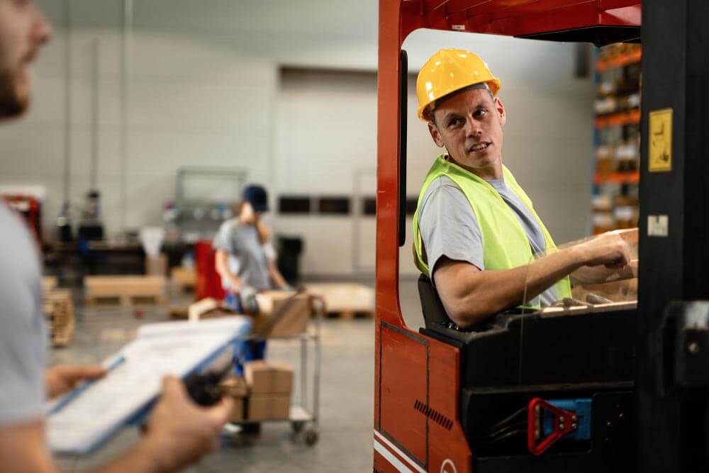 forklift operator, wearing a hard hat and a safety vest, is in conversation with their supervisor